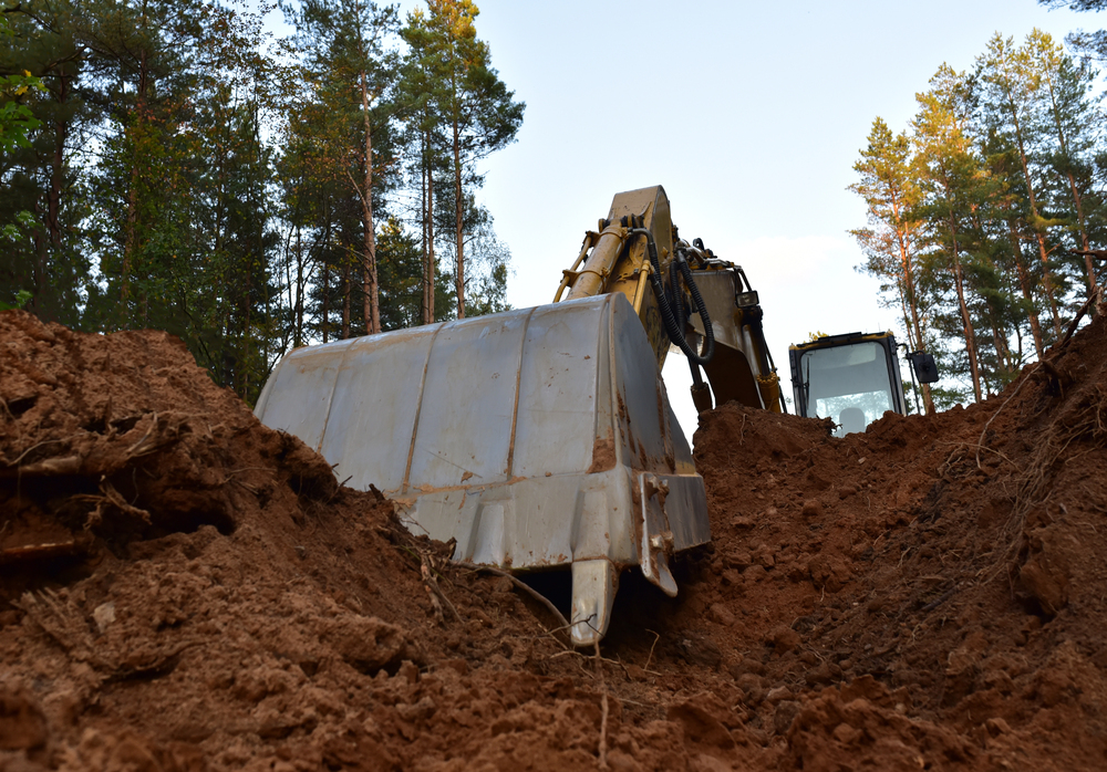 A large excavator is digging in the dirt at a construction site surrounded by trees, with its bucket raised and filled with soil.