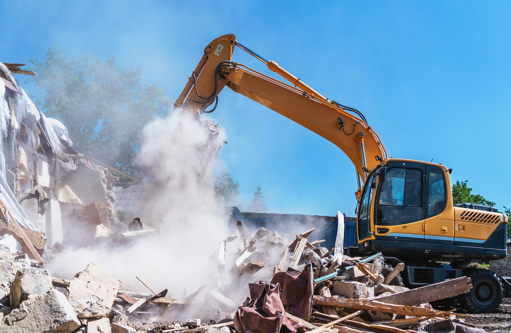 An excavator demolishes a building, creating a cloud of dust amid piles of debris under a clear blue sky.