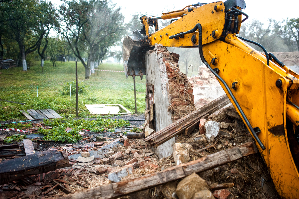 A yellow excavator demolishes a brick wall in an outdoor area, with debris and rubble scattered on the ground. Trees and grass are visible in the background.