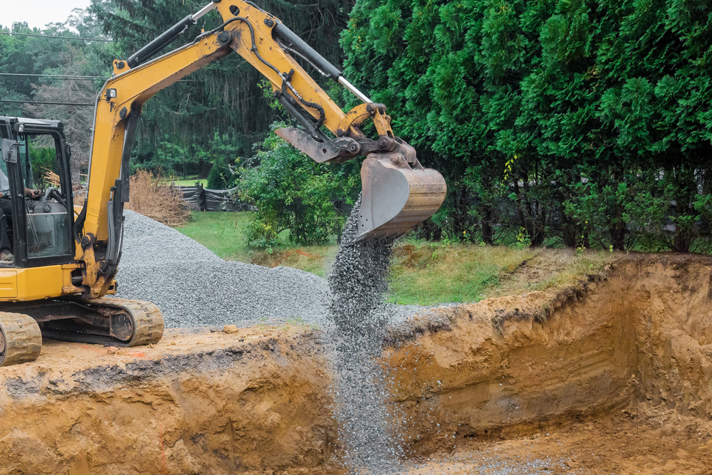 A yellow excavator pours gravel into a dug-out area at a construction site bordered by green trees.