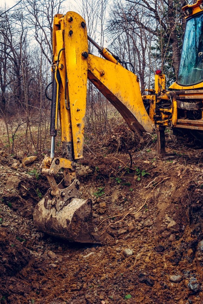 A yellow excavator with its bucket digging into a patch of earth in an outdoor, wooded area.