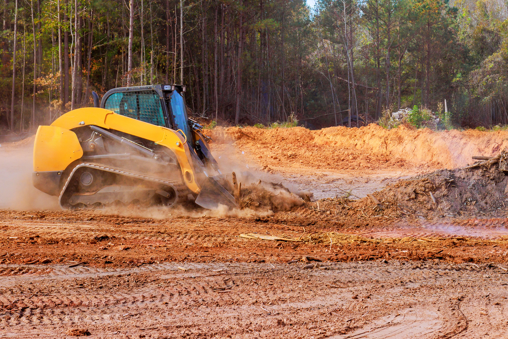 A yellow skid steer loader moves dirt and debris on a construction site surrounded by trees on a sunny day.