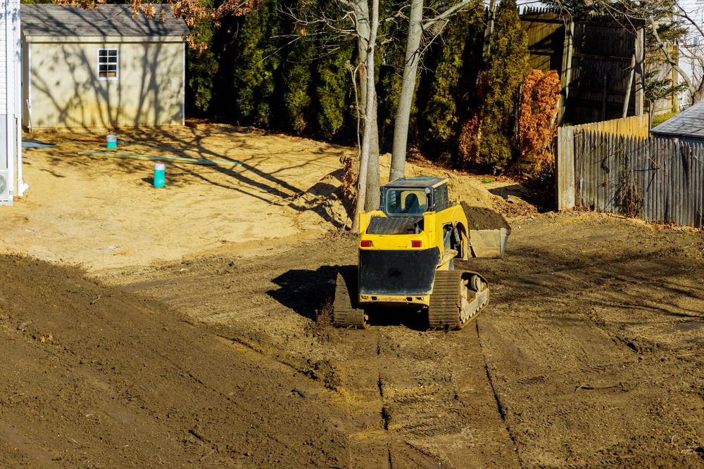 A yellow bulldozer is grading soil in a yard near a tan shed, with trees, a wooden fence, and some exposed blue pipes in the background.