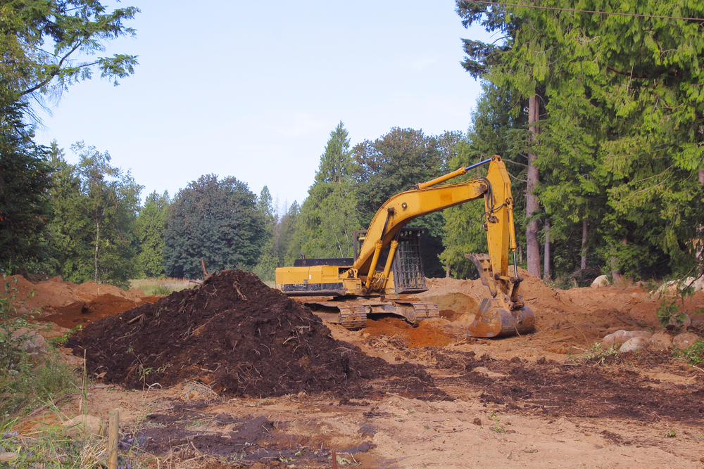 A yellow excavator is parked on a dirt construction site, surrounded by piles of soil and trees in the background.
