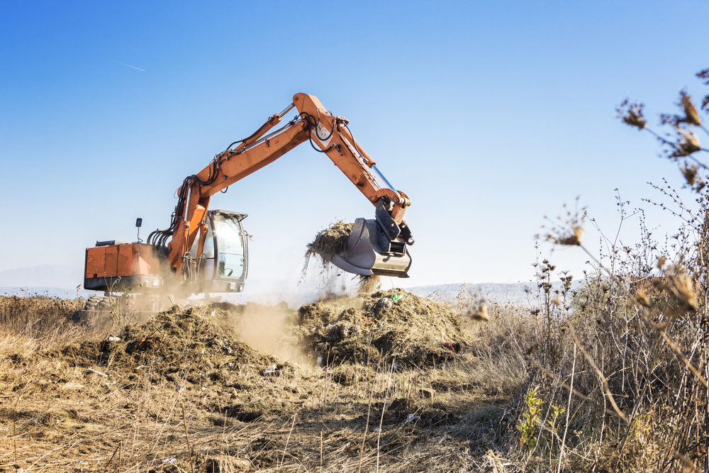 An orange excavator moves a pile of dirt and debris on a dry, grassy field under a clear blue sky.